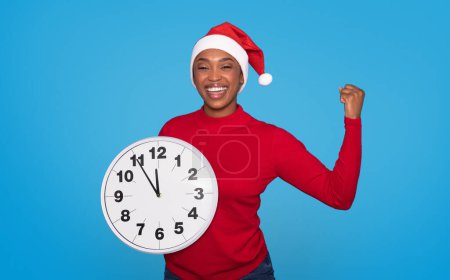 Téléchargez les photos : Femme afro-américaine joyeuse dans Santa Hat tenant une grande horloge sur fond bleu en studio, montrant compte à rebours pour la nouvelle année à venir, geste oui et sourire à la caméra - en image libre de droit