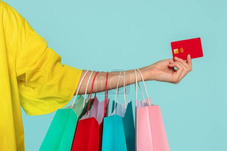 Foto de Cultivado de mujer sosteniendo compras coloridas bolsas de compras y tarjeta de crédito bancaria de plástico rojo, aislado en fondo azul estudio. Compras ilimitadas sin contacto, primer plano - Imagen libre de derechos