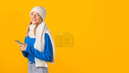 Foto de Mujer alegre en suéter azul, bufanda blanca y sombrero, se involucra con su teléfono inteligente, sugiriendo momento de comunicación agradable, sobre fondo amarillo, panorama - Imagen libre de derechos