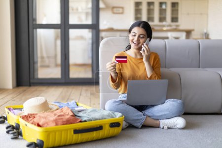 Photo for Happy young indian woman with credit card and cellphone booking vacation at travel agency or making hotel reservation online, using laptop computer, planning trip, sitting next to suitcase at home - Royalty Free Image