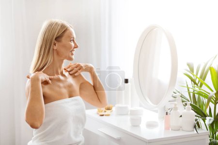 Photo for Admiring Beauty. Attractive Middle Aged Woman Wrapped In Towel Sitting At Dressing Table At Home, Happy Mature Female Looking In Mirror And Smiling, Enjoying Domestic Self-Care Routine, Copy Space - Royalty Free Image