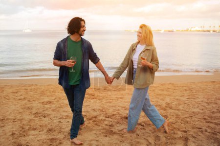 Photo for Young european couple sipping wine while walking on the beach, affectionate man and woman holding hands and gazing at each other, happy lovers enjoying romantic date outdoors, copy space - Royalty Free Image
