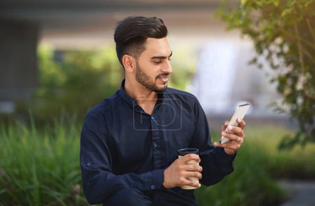 Téléchargez les photos : Homme d'affaires arabe souriant utilisant le téléphone et communiquant via des messages en ligne dans l'application de médias sociaux, assis sur un fond de rue verte animée dans la zone urbaine. gadgets modernes - en image libre de droit