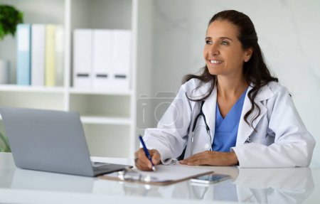 Photo for Smiling mature woman doctor taking notes in medical chart, sitting at workdesk with laptop at her cabinet in clinic, taking anamnesis while having conversation with patient, looking at free space - Royalty Free Image