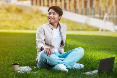 Photo for Cheerful asian man student using laptop and learning online, doing homework outdoors, sitting on lawn in university campus park. Guy studying distantly and smiling. Freelancer working online - Royalty Free Image