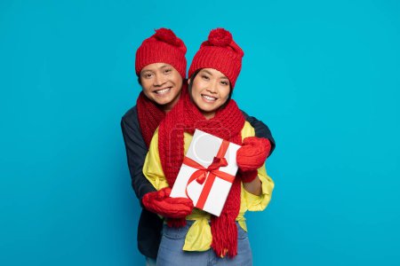 Photo for Happy millennial asian guy in hat show box present and hugs woman, isolated on blue studio background. Weekend, cold, holidays, winter and New Year celebration together, Xmas gifts - Royalty Free Image