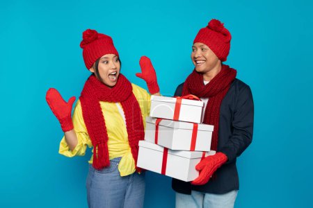 Téléchargez les photos : Souriant jeune homme asiatique en chapeau donne de nombreuses boîtes présentes à dame surprise, isolé sur fond de studio bleu. Couple profiter week-end, vacances, hiver et Nouvel An, cadeaux de Noël - en image libre de droit