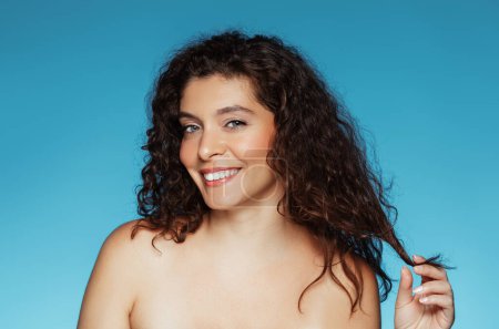 Photo for Glad young caucasian curly naked lady touching hair, enjoy beauty care result, isolated on blue studio background. Wellness, treatment and spa procedures, lifestyle, sale - Royalty Free Image
