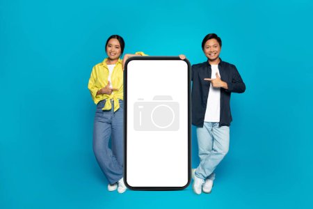 Photo for Happy young asian couple in casual pointing fingers at big smartphone with empty space, isolated on blue studio background. Recommendation gadget, presentation, ad and offer - Royalty Free Image