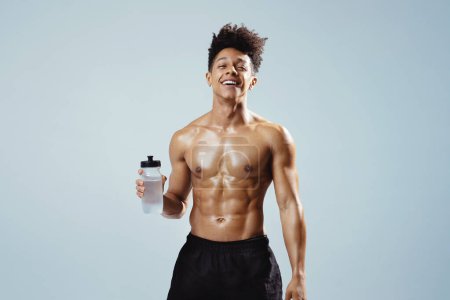 Photo for Workout Hydration. Cheerful Athletic Young Man Holds Bottle Of Water And Smiles To Camera On Gray Background. Studio Shot Of Handsome Muscular Fitness Guy With Shirtless Torso Hydrating After Sport - Royalty Free Image