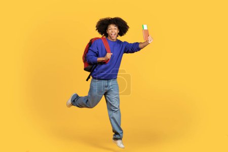 Téléchargez les photos : Heureux voyageur. Portrait de sourire homme noir avec sac à dos, passeport et billets de voyage sautant sur fond jaune studio, joyeux jeune homme afro-américain prêt pour le voyage de vacances - en image libre de droit