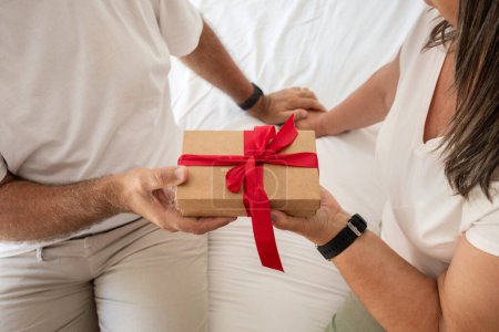 Photo for Positive senior caucasian woman gives gift box to man, sit on bad in bedroom interior, close up. Celebration birthday, anniversary surprise, holiday together, love and congratulate at home - Royalty Free Image