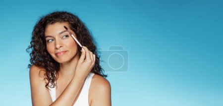 Téléchargez les photos : Femme bouclée européenne millénaire positive avec brosse peignant les sourcils, fait maquillage de base, isolé sur fond bleu studio, panorama. Soins de beauté et routines quotidiennes, mode - en image libre de droit