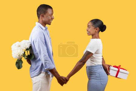 Photo for Black couple with flowers and gift celebrating lovers holiday, sweethearts hiding surprises for each other behind their backs over yellow studio background, side view - Royalty Free Image