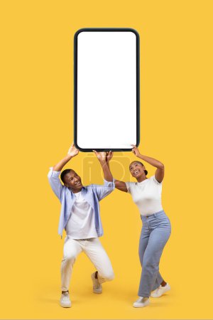 Téléchargez les photos : Couple noir tenant un énorme smartphone lourd avec écran noir blanc sur fond de studio jaune, maquette pour l'application mobile ou la publicité sur le site Web, grande offre - en image libre de droit