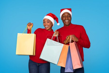 Téléchargez les photos : Saison de Noël. Happy Black Spouses Publicité vacances Shopping offre, tenant de nombreux sacs de papier Shopper sur fond bleu studio. Acheteurs posant dans Santa Chapeaux, Souriant à la caméra - en image libre de droit