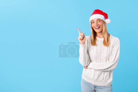 Photo for Happy woman in white knit sweater and Santa hat points upwards at free space, creating celebratory atmosphere perfect for the holiday season, blue background, banner - Royalty Free Image