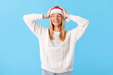 Foto de Joven alegre, vistiendo suéter blanco y sombrero de Santa, celebra la temporada festiva, sonriendo con las manos en la cabeza, exudando el espíritu jubiloso de la Navidad sobre fondo azul - Imagen libre de derechos