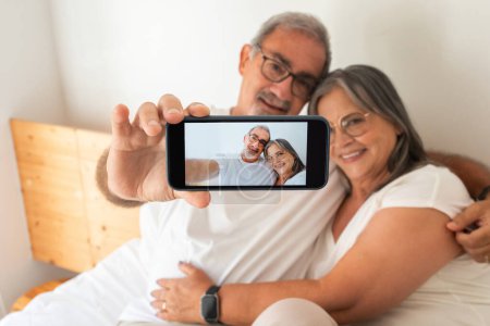 Photo for Happy old european husband and wife make selfie on phone screen on bed in bedroom interior. Rest and relaxation together, photo app and device for social networks at spare time at home - Royalty Free Image