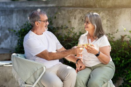 Téléchargez les photos : Positif homme caucasien senior et dame acclamations verres de vin assis sur des chaises dans le jardin à la maison. Rendez-vous romantique, célébration des vacances, relation, amour et temps libre ensemble - en image libre de droit