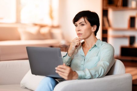 Photo for Thoughtful middle aged European woman with laptop pondering on a business concept, websurfing on her gadget, lounging on couch in living room indoor. Weekend of productive thought - Royalty Free Image