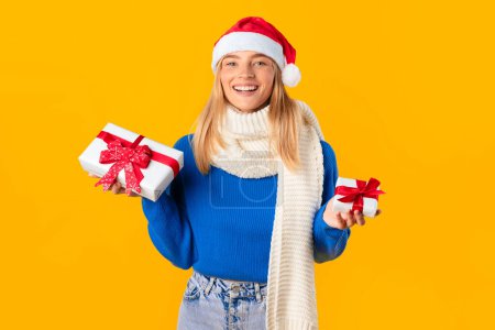 Photo for Cheerful woman in festive Santa hat excitedly presents wrapped Christmas gifts with bright red ribbons, embodying the joy of winter Christmas sales, against vibrant yellow backdrop - Royalty Free Image