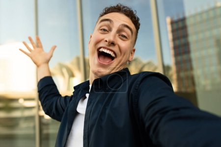 Téléchargez les photos : Heureux blogueur homme caucasien millénaire excité avec la bouche ouverte, profiter du mode de vie, faire du selfie au téléphone dans la ville, près de l'immeuble de bureaux. Travail, étude et photo pour blog, réseaux sociaux - en image libre de droit