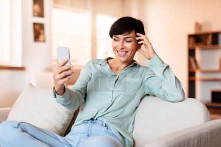 Téléchargez les photos : Femme brune d'âge moyen souriante naviguant sur les médias sociaux sur son téléphone portable, exprimant son bonheur tout en se prélassant sur le canapé dans le salon à la maison. Loisirs et communications numériques - en image libre de droit