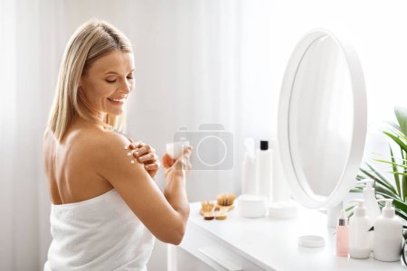 Photo for Skin Nourishing. Beautiful Smiling Middle Aged Woman Applying Body Cream, Portrait Of Attractive Mature Lady Wrapped In Towel Using Moisturising Lotion After Bath, Sitting At Dressing Table At Home - Royalty Free Image