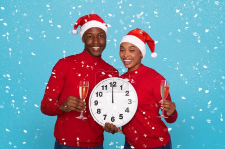 Photo for Happy African American Couple In Santa Hats Holding Champagne Glasses And Large Clock Amidst Falling Confetti On Blue Studio Background, Waiting Midnight Celebration And New Years Eve Festivities - Royalty Free Image