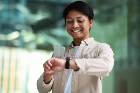 Téléchargez les photos : Souriant asiatique millénaire homme vérifie le temps sur smartwatch utilise l'application pour surveiller le temps habillé en tenue décontractée pose à l'extérieur sur fond flou, tenant smartphone dans sa main - en image libre de droit