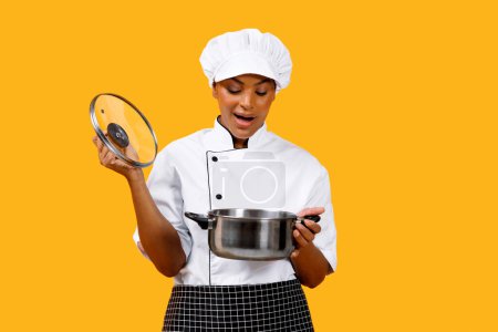 Photo for Surprised African American Chef Lady Looking Into Saucepan, Excited Black Cook Woman Holding Stewpot In Hands And Checking Meal While Cooking, Posing Isolated On Yellow Studio Background, Copy Space - Royalty Free Image