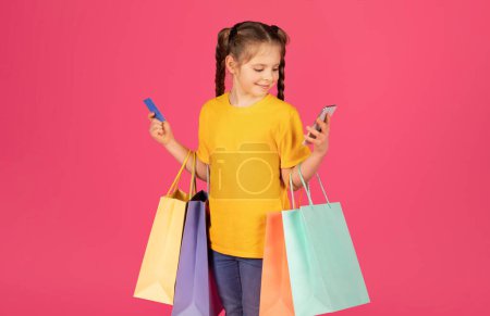 Foto de Retrato de linda niña preadolescente sosteniendo tarjetas de crédito y bolsas de compras y el uso de Smartphone, Sonriente niña aplicación de navegación en el teléfono móvil para compras en línea, mientras que de pie sobre fondo rosa - Imagen libre de derechos
