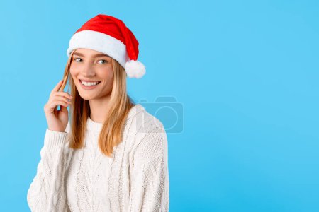 Téléchargez les photos : Heureuse jeune femme européenne au chapeau de Père Noël touche ses cheveux, jetant un regard curieux sur le côté sur un fond bleu d'hiver, capturant une humeur festive. - en image libre de droit