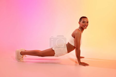 Foto de La joven latina positiva en ropa deportiva hace flexiones, entrenamiento de tablón en el suelo sobre fondo de estudio de color rosa neón. Fitness, deporte, cuidado de la salud y pérdida de peso, ejercicios solos - Imagen libre de derechos