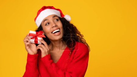 Foto de Encantada dama negra con sombrero de Santa y suéter rojo sostiene regalo envuelto con cinta roja, irradiando el espíritu de las vacaciones de invierno sobre el fondo amarillo, espacio de copia - Imagen libre de derechos