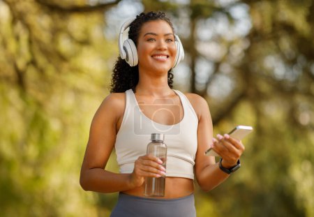 Photo for Cheerful millennial brazilian lady athlete in sportswear with wireless headphones holding phone and fitness bottle, listening to music during morning jog, exercising in green park outside - Royalty Free Image
