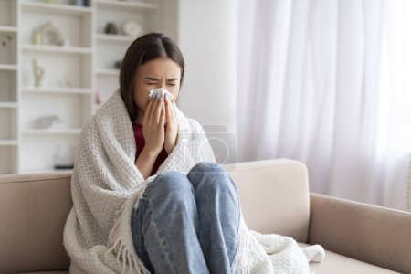 Foto de Mujer asiática enferma joven que sopla la nariz en el tejido de papel mientras está sentada en el sofá en casa cubierta en manta, retrato de la enferma mujer coreana que sufre secreción nasal, sensación de malestar, tener gripe estacional - Imagen libre de derechos