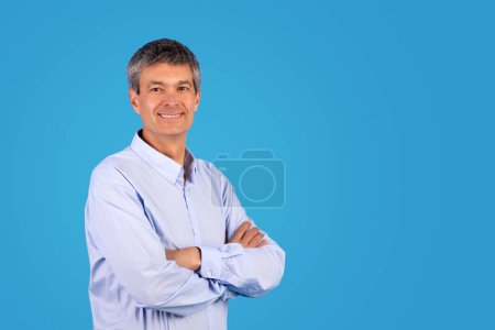 Foto de Hombre maduro positivo en camisa azul casual posando con confianza contra el telón de fondo del estudio, cruzando las manos mientras está de pie, promoviendo la oferta con sonrisa, espacio en blanco para la publicidad - Imagen libre de derechos