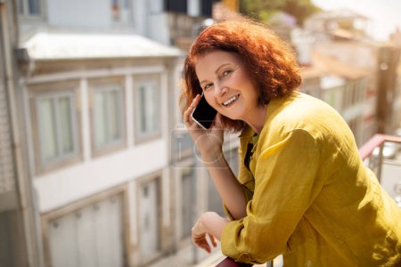 Foto de Mujer mayor sonriente hablando en el teléfono celular mientras está de pie en el balcón en casa, feliz pelirroja anciana teniendo agradable conversación telefónica y disfrutando de la vista, mirando a la cámara, Copiar el espacio - Imagen libre de derechos