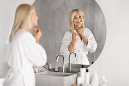 Photo for Split Ends Treatment. Beautiful Mature Woman Applying Serum On Damaged Dry Hair While Standing Near Mirror In Bathroom, Happy Senior Female Doing Haircare Routine, Selective Focus On Reflection - Royalty Free Image
