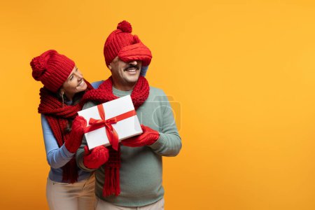 Foto de Sonriente esposa europea senior en sombrero cierra los ojos con la mano al marido en invierno, caja de regalo presente, aislado en fondo de estudio naranja. Las relaciones, el regalo para Nuevo Año, la celebración de Navidad - Imagen libre de derechos