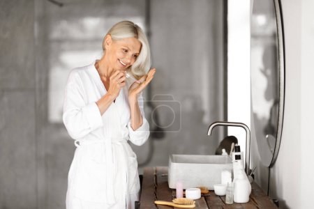 Photo for Haircare routine. Smiling senior woman applying hair serum on her dry ends, beautiful mature female in bathroom making beauty treatments while standing near mirror in bathroom, copy space - Royalty Free Image