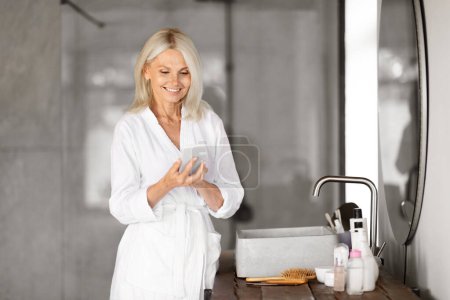 Smiling Senior Woman With Smartphone Reading Beauty Hacks While Getting Ready Near Mirror In Bathroom, Attractive Mature Female Browsing Internet On Mobile Phone, Shopping Cosmetics Online
