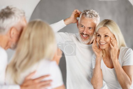Téléchargez les photos : Concept de soins de beauté. Heureux couple âgé faisant routine du matin dans la salle de bain, joyeux conjoints matures souriant à leur reflet dans le miroir, Femme appliquant crème visage, Focus sélectif - en image libre de droit