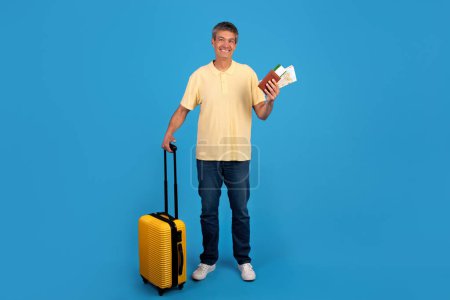 Téléchargez les photos : Globetrotter gris homme avec passeport et billets à la main, prêt pour les vacances d'été, plan complet du voyageur avec valise debout sur fond bleu. Joie du voyage de vacances, annonce d'agence de voyage - en image libre de droit