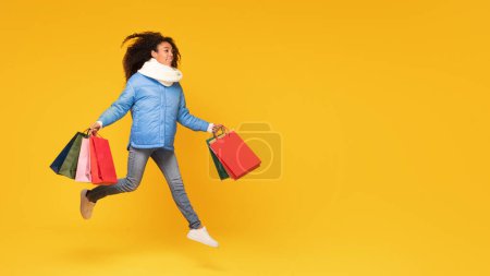 Foto de Feliz dama negra vestida con moda de invierno sostiene coloridas bolsas de compras, mostrando la emoción de las ventas de invierno y promociones contra el fondo amarillo, panorama, espacio libre - Imagen libre de derechos