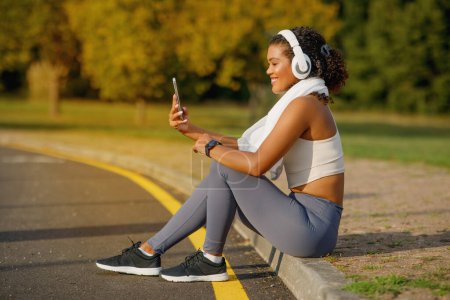 Téléchargez les photos : Vue latérale de l'athlète brésilienne avec écouteurs vidéo appelant sur smartphone, repos de l'entraînement assis en plein air, l'exercice dans le parc public le matin. Application sportive et soins du corps - en image libre de droit