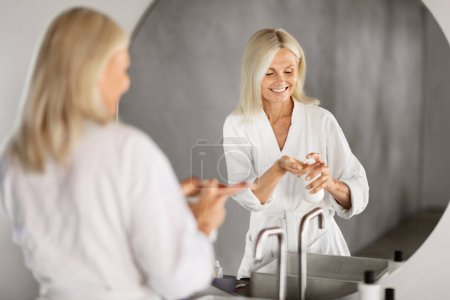 Téléchargez les photos : Belle femme âgée utilisant une lotion hydratante tout en se tenant près du miroir dans la salle de bain, souriante jolie dame mature en peignoir tenant bouteille avec des produits de beauté, profiter de la routine d'auto-soins - en image libre de droit