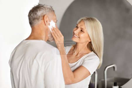 Photo for Loving Mature Wife Applying Shaving Foam On Husbands Face In Bathroom, Happy Senior Couple Having Fun While Getting Ready In Morning Together, Making Daily Beauty Routine At Home, Copy Space - Royalty Free Image